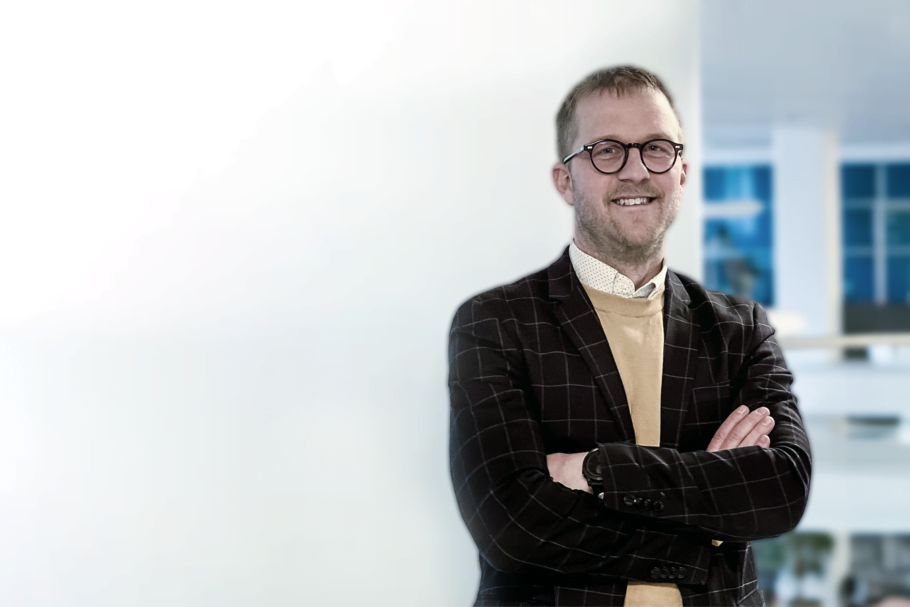 Läraren Henrik Arnersten, iförd rutig kavaj, beige långärmad, vit skjorta och runda glasögon, står med armarna i kors framför en vit vägg i en av skolans korridorer och ler mot kameran.