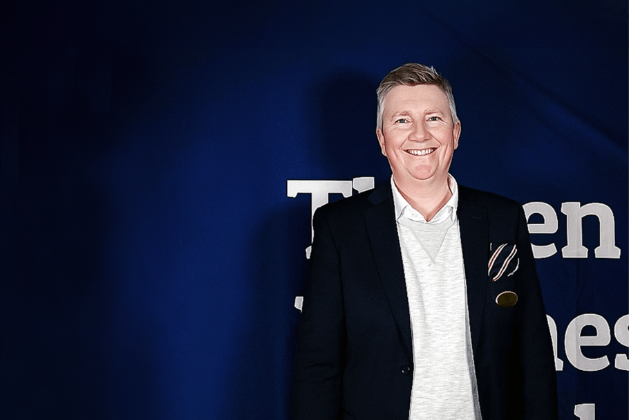 Läraren Andreas Liljeson, iförd marinblå kavaj och grå långärmad tröja över en vit skjorta, står framför en mörkblå bakgrund och ler brett mot kameran.