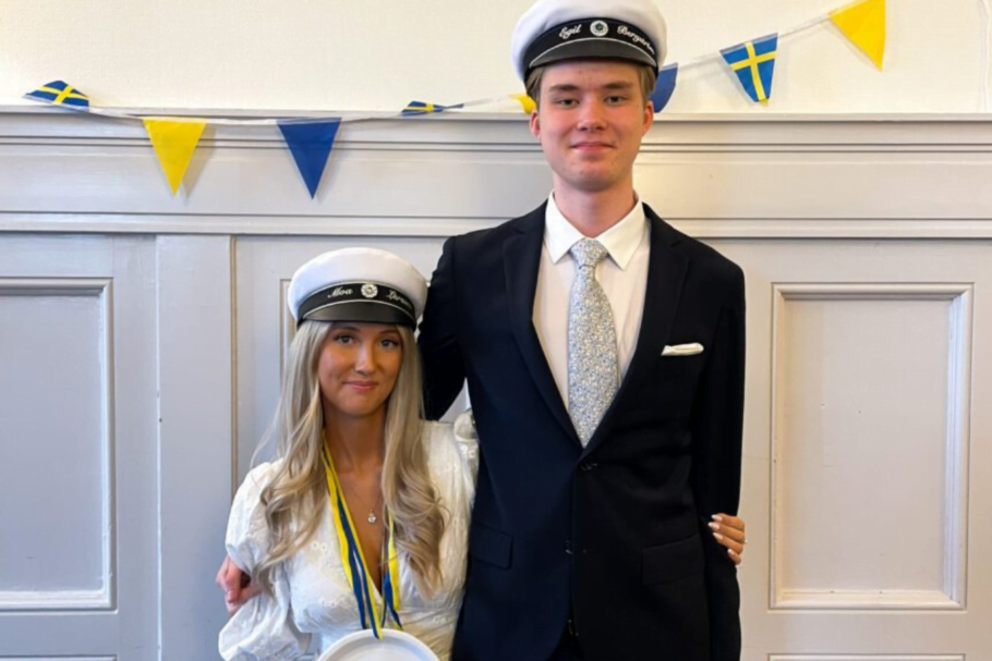 Moa Larssen och Egil Bergström på Thoren Business School Gävle firar studenten. Moa är klädd i vit klänning och studentmössa, Egil har kostym, även han med studentmössa. Bakgrunden pryds av svenska flaggor och vimplar.