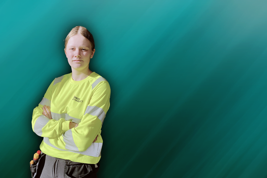 Yrkesgymnasiet-eleven Nora, iförd en guld varseltröja och mörka arbetsböcker, står med armarna i kors och ler mot kameran framför en grönmelerad fondvägg.