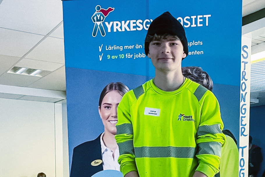 Eleven Oskar Hennström står iförd Yrkesgymnasiets gula varseltröja och en svart mössa framför en blå roll-up med information om skolan. Han står med händerna knäppta framför midjan och ler mot kameran.