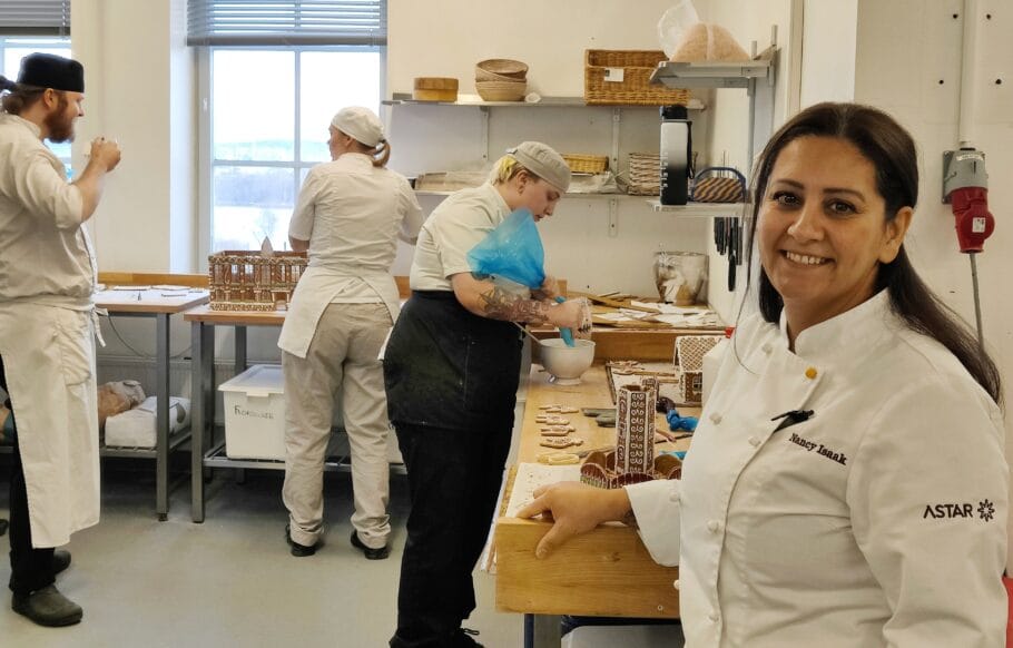 En kvinnlig lärare i vit kockrock med texten 'Nancy Isaak' och 'Astar' på ärmen står vid ett arbetsbord i ett bagerikök. Bakom henne arbetar elever i vita arbetskläder med att dekorera pepparkakshus och kakor, medan en elev spritsar glasyr.