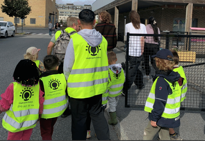 En grupp barn och vuxna går tillsammans utomhus. Barnen bär gula reflexvästar med texten "Långbroparks förskola" och ett telefonnummer på ryggen. De leds över en gata mot en byggnad, och några vuxna följer med som ledare.