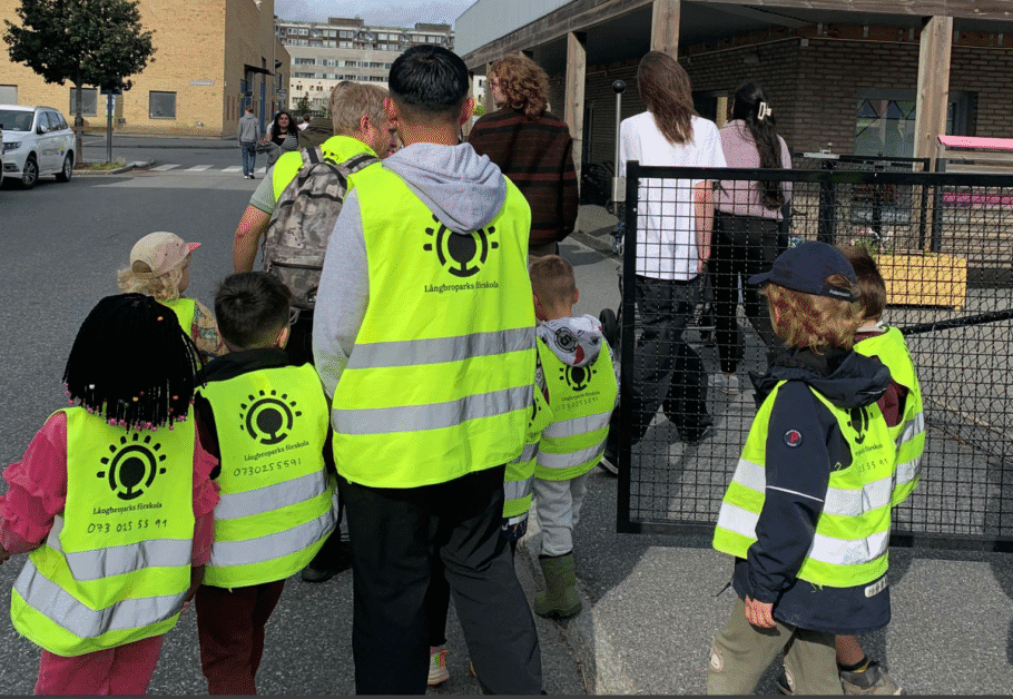 En grupp barn och vuxna går tillsammans utomhus. Barnen bär gula reflexvästar med texten "Långbroparks förskola" och ett telefonnummer på ryggen. De leds över en gata mot en byggnad, och några vuxna följer med som ledare.