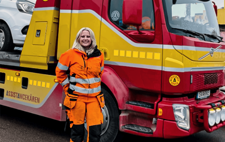 Eleven Emmy står i orange arbetskläder och ler framför en röd bärgningsbil.