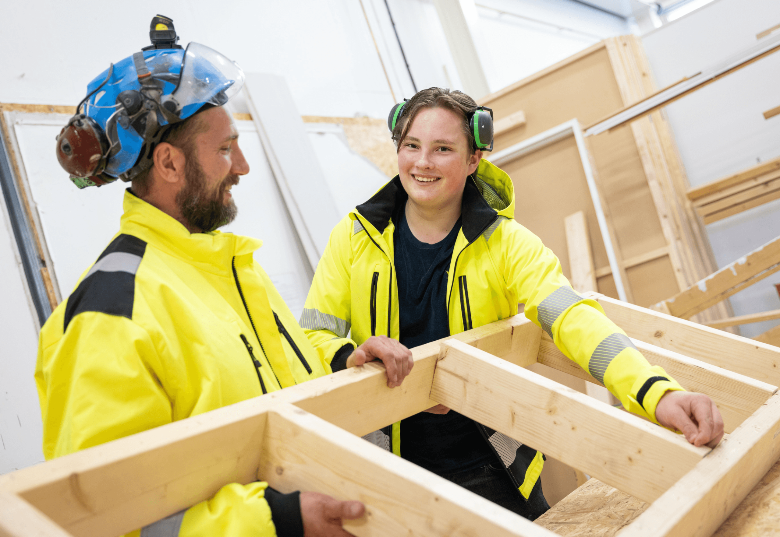 En elev och en lärare iklädda varselkläder och hörselkåpor lyfter en träram på en byggplats.