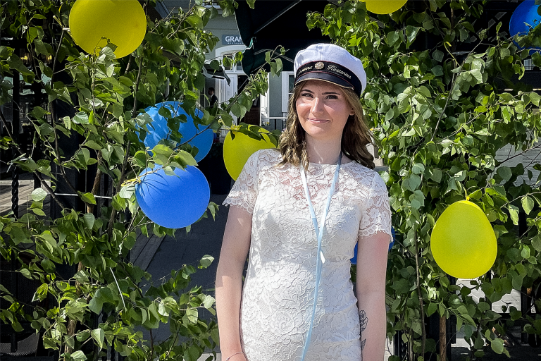 En leende och stolt Savanna Kostenius iförd full studentmundering framför gröna buskar dekorerade med gula och blå balkonger under en solig, svensk sommardag.