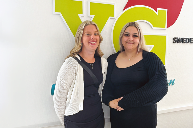 Eleverna Nathalie Strömquist Ana Iljazovic och står och ler mot kameran, framför en vit vägg med Yrkesgymnasiets logotyp.