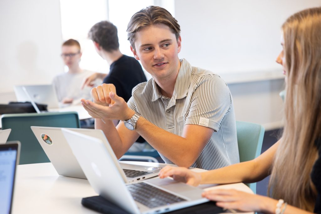 Gymnasieelever som samarbetar vid datorer i klassrum.