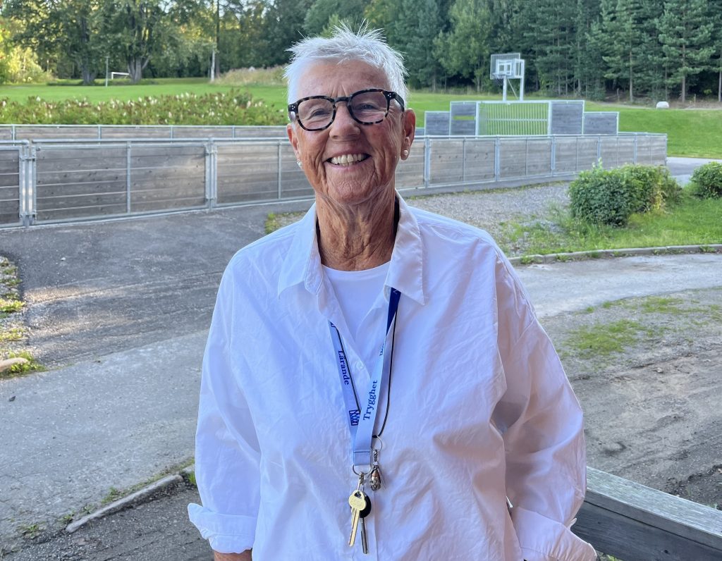 Ett foto på Lena Wallgren utanför skolan där man ser skolgården med gräs och skog.