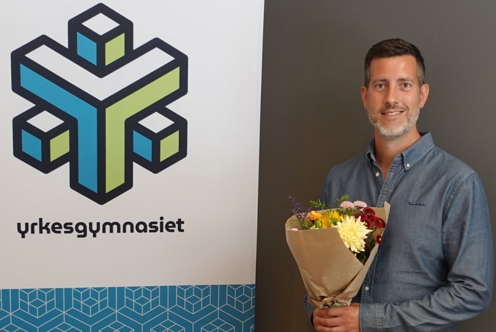 Marcos Améztegui med en blomsterbukett i handen och bredvid en roll-up med Yrkesgymnasiets logga på.