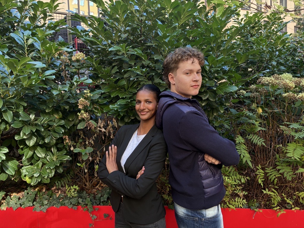De står med rygg mot rygg framför en låg röd mur med buskage ovan.